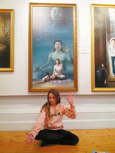 圖5：澳洲女孩何莉坐在《純凈入仙境》畫作前，模仿著畫中女修煉者的姿勢打手印，家人很高興，一家人表示要去煉功點學功。