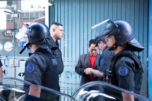 '图中正在被警察扣押，盘问、身穿红色体恤深色外套的男子是在二零零四年罗干到阿根廷时，殴打和平抗议罗干的法轮功学员的报道，并因此而被阿根廷法院通缉的疑犯。'