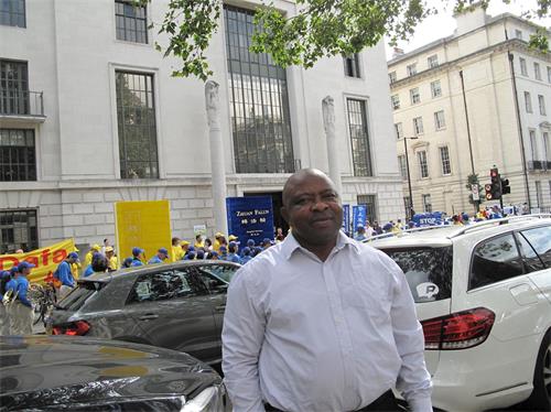 '图10：八月三十日，在伦敦中使馆前，在伦敦银行工作的西尔维塞尔（Sylveseer）驻足聆听并拍摄法轮功学员的新闻发布会'