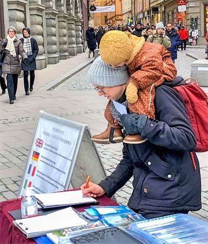 '图5：这位瑞典人在签名支持制止迫害法轮功时，骑在爸爸肩上的宝宝认真地注视着爸爸签名。'