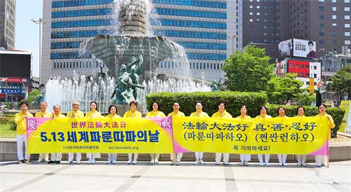 图1：5月13日，部分首尔西部和中部的法轮功学员们在首尔市中心新世界百货店附近的大型喷泉前举行了集体炼功、派发传单等洪法活动。图为他们在活动前，首先恭祝慈悲伟大的师尊华诞。